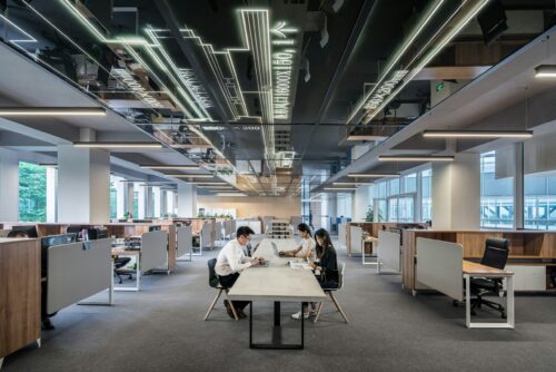 Grand open space de bureau moderne avec plusieurs postes de travail, personnes en réunion autour d’une table centrale.